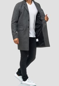 Manteau gris en mélange de laine avec une fermeture à un bouton, deux poches avant et une doublure intérieure lisse, porté sur une chemise blanche et un jean noir.