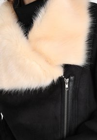 Giacca in suede nera con colletto in pelliccia sintetica di un morbido crema, con chiusura frontale a zip completa e una tasca laterale con zip angolata.