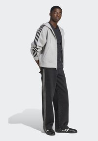 Šedá mikina na zips s čiernymi pruhmi pozdĺž rukávov, v kombinácii s čiernymi širokými nohavicami s šedými bočnými pruhmi, doplnená čiernymi teniskami.