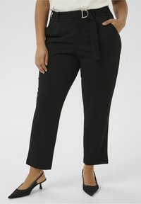 Femme portant un pantalon noir ajusté avec une ceinture en tissu et des escarpins à talons hauts noirs à bout pointu, se tenant avec une main dans la poche.