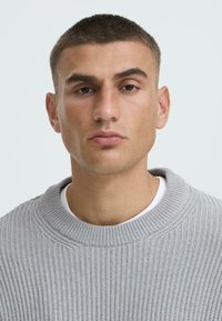 Pelēks ribots džemperis ar platu apkakli, uzvilkts virs baltas krekla. Tuvplāns uz personu ar īsiem matiem un neitrālu izteiksmi.