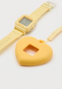 Κίτρινο ψηφιακό ρολόι χειρός με ορθογώνιο καντράν δίπλα σε κίτρινη προστατευτική θήκη σε σχήμα καρδιάς με ιμάντα χειρός, και τα δύο με την ένδειξη BABY-G.