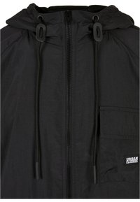 Black lightweight jacket featuring a zip front, adjustable hood with toggle, and a side pocket with a logo detail. Textured fabric.