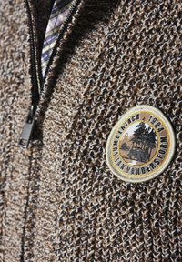 Brauner Strick-Übergangspullover mit strukturiertem Muster und einem runden Patch mit einem Baum- und Bergdesign, beschriftet mit "Lumberjack 1984."