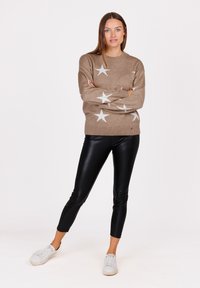Beige gestrickter Pullover mit weißem Sternmuster, Rundhalsausschnitt und langen Ärmeln, kombiniert mit glänzenden schwarzen Leggings und weißen Turnschuhen.