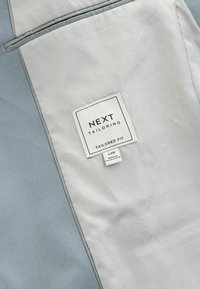 Maßgeschneiderte Anzugjacke Innenfutter, hellgraue Fütterung mit subtilen Mustern, blaue Außenstoff, Etikett mit Marken- und Größenangaben.