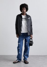 Zwarte button-up jas over een witte graphic T-shirt, gecombineerd met versleten blauwe jeans met metallic details en zwarte sandalen. Inclusief een kleine handtas.