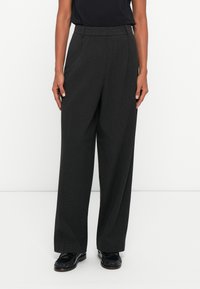 Calças largas, de cintura alta, cinzentas, com um tecido texturizado, apresentando pregas frontais e bolsos laterais. Combinadas com sapatos pretos.