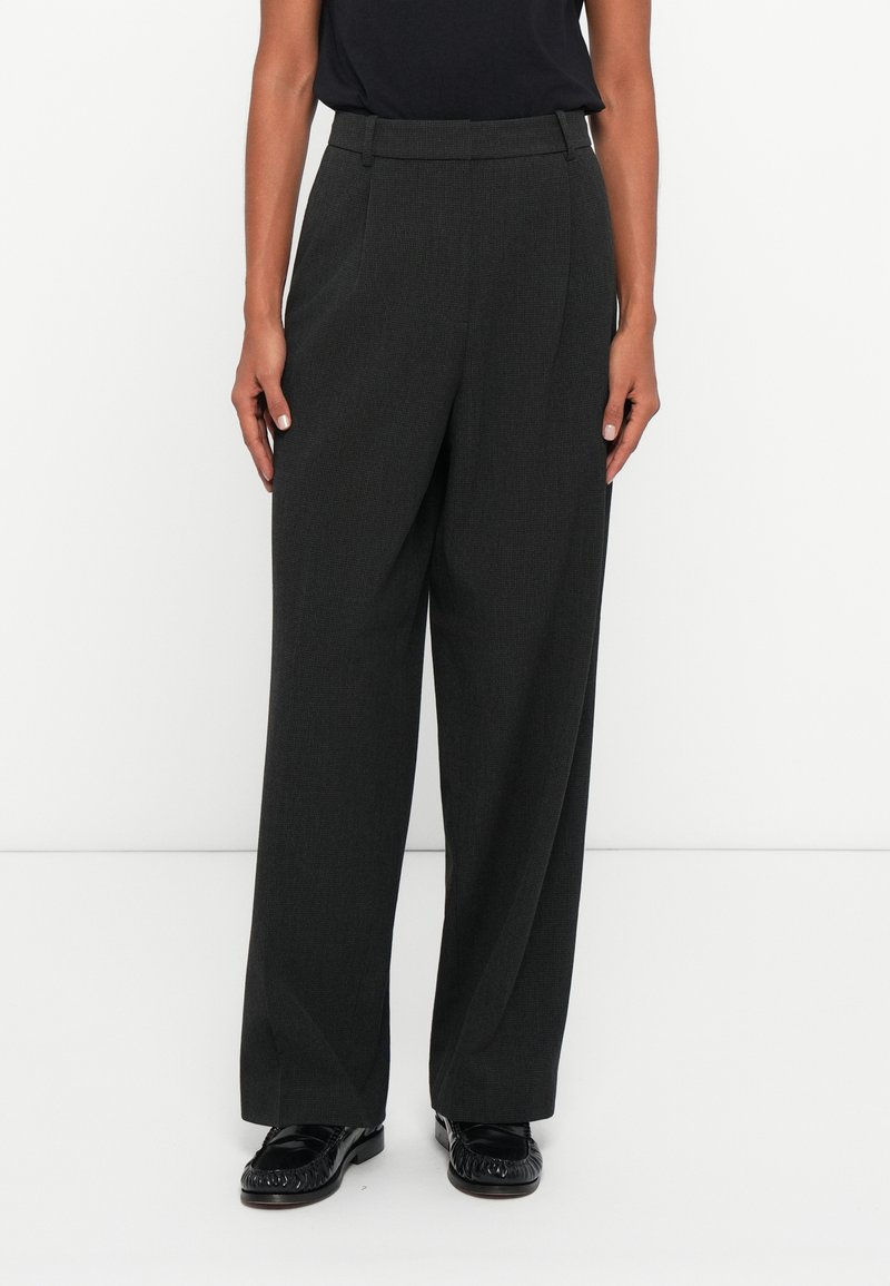 Calças largas, de cintura alta, cinzentas, com um tecido texturizado, apresentando pregas frontais e bolsos laterais. Combinadas com sapatos pretos.