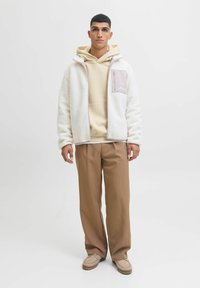White fleece jacket with a beige zip pocket, paired with a tan hoodie and brown wide-leg trousers, complemented by beige moccasin shoes.