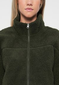 Giacca in fleece verde oliva con colletto alto, chiusura frontale con zip e tessuto testurizzato caratterizzato da una fodera interna liscia e priva di motivi.