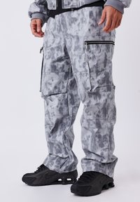 Grijze camouflage cargo-broek met meerdere zakken en ritsaccenten, gemaakt van een zacht materiaal. Gemaakt met zwarte sport schoenen.