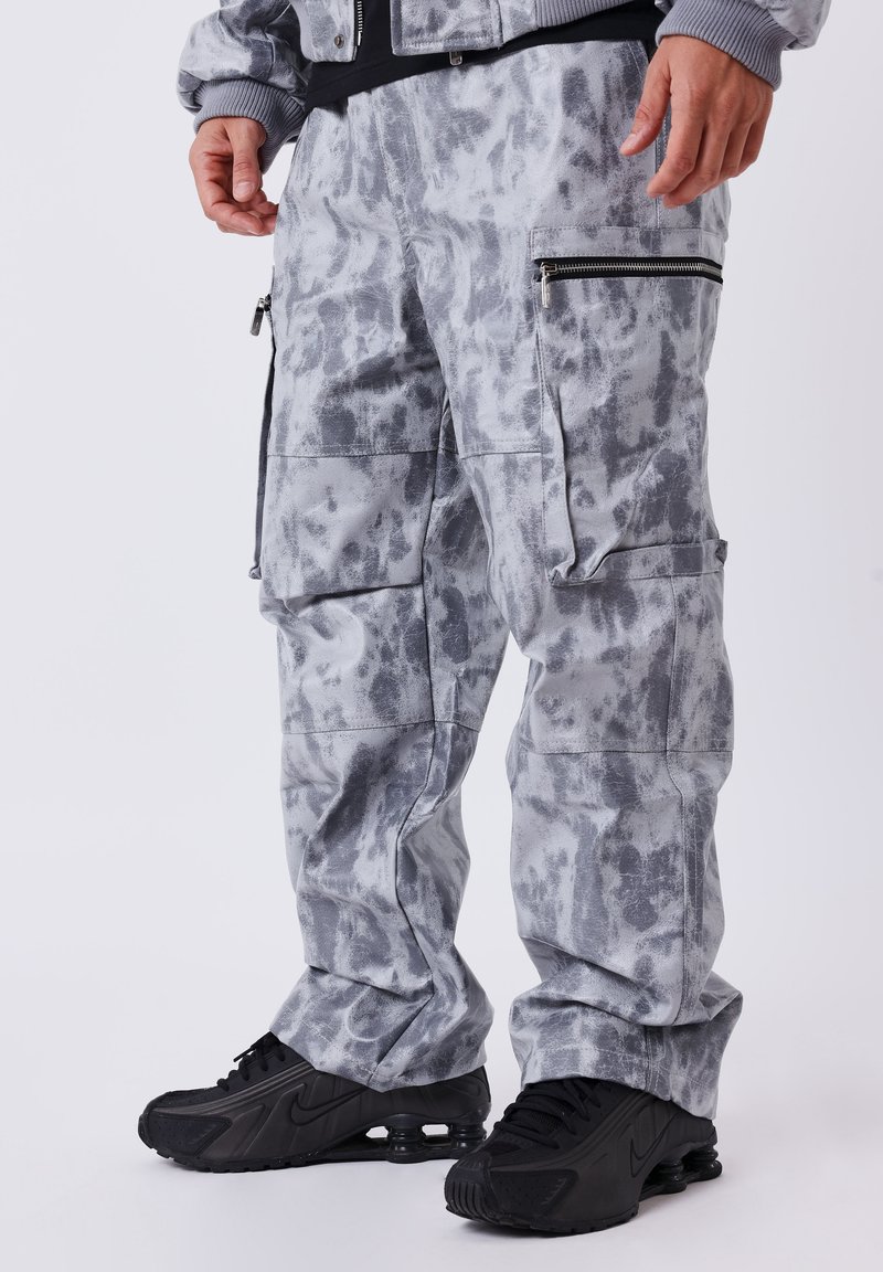 Grijze camouflage cargo-broek met meerdere zakken en ritsaccenten, gemaakt van een zacht materiaal. Gemaakt met zwarte sport schoenen.