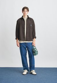 Camicia a quadri marrone con bottoni, t-shirt beige chiaro, pantaloni in denim blu, sneakers bianche e una piccola borsa verde. Il modello si trova su un tappeto blu.