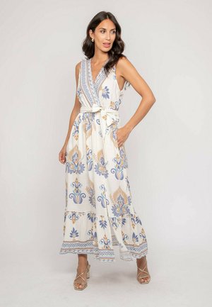 Femme portant une robe longue blanche sans manches avec des motifs floraux bleus et beige, ceinture nouée à la taille, et sandales beiges à talons, debout avec la main dans la poche.