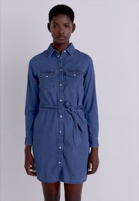 Denim skjortekjole i mørkeblå, med kragehals, lange ermer, to brystlommer og en belte som knytes i livet.