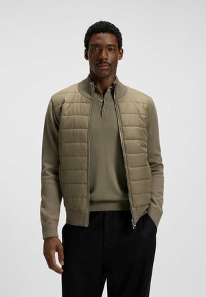 Giacca imbottita verde oliva con zip frontale, collo a costine e sezioni in morbido tessuto a maglia. Indossata sopra una polo abbinata e pantaloni scuri.