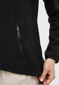 Veste en polaire noire avec une poche zippée, un cordon élastique à l'ourlet et une texture brossée pour chaleur et confort.