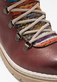 Bota de cuero marrón con parte superior de textil multicolor trenzado, cordones beige y ojetes de metal. Presenta una suela de goma blanca texturizada.