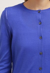 Pletený modrý kardigan s kulatým výstřihem, pěti černými knoflíky a jemnou texturou s žebrovaným panelem na rameni.