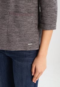 Szary dzianinowy top z teksturowanym wzorem, okrągłym dekoltem i podwiniętymi rękawami do połowy ramienia. Posiada przednią kieszeń z detalicznym przeszyciem oraz metkę z logo.