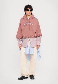 Felpa cropped in rosa polveroso oversize con logo "AVAVAV", orlo sfrangiato, sovrapposta a una camicia a righe blu e bianca, abbinata a pantaloni crema.
