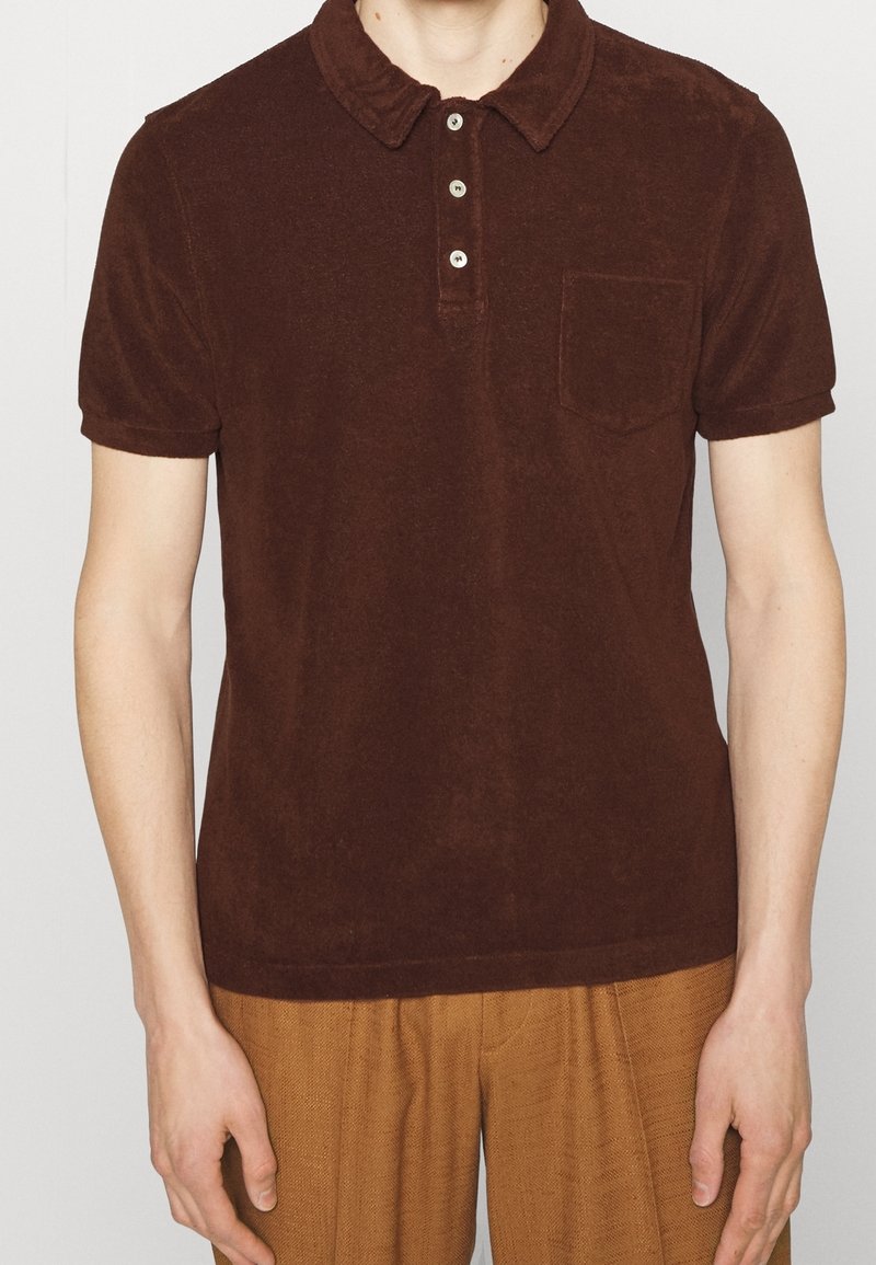 Personne portant un polo marron foncé à manches courtes avec trois boutons et une poche poitrine, associé à un pantalon marron clair.