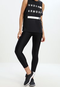 Osoba oblečená v černém bezrukávovém triku Under Armour, černých legínách do kotníků a černých sportovních botách s bílou podrážkou, stojící s překříženýma nohama.