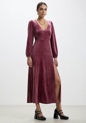 Robe midi en velours de couleur rose foncé avec de longues manches ballon, un décolleté en V plongeant, des détails de boutons et une fente sur le côté. Associée à des chaussures à talons noires.