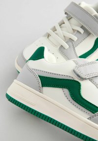 Hvide og grå sneakers med grønne detaljer, der har en glat læderoverdel, tekstureret sidepanel, flade snørebånd og en Velcrorem.