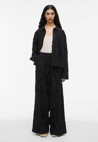 Frau mit langen dunklen Haaren, die ein schwarzes, floral gemustertes Oversize-Hemd und weit geschnittene Hosen, ein weißes Tanktop und beigefarbene Schuhe mit quadrater Zehenform trägt.