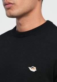 Svart strikket genser med rund hals og ribbestrikkede mansjetter, med et lite brodert merkelapp på brystet.