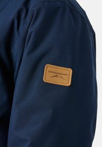 Marineblauwe jas met een gestructureerde stof en een tan leren patch met "SWEDEMOUNT" en een bergafbeelding op de bovenarm.