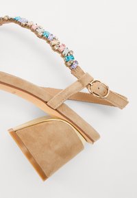 Beige semsket sandal med en kraftig gullbelagt hæl. Har en stropp pyntet med flerfargede rhinestones og et justerbart spenne.