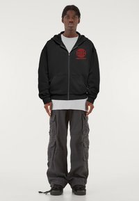 Fekete cipzáras kapucnis pulóver piros grafikával és felirattal a mellkason, szürke hosszú ujjú pólóval és szürke cargo nadrággal párosítva, fekete cipővel viselve.