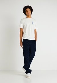 Tricou alb din bumbac cu mâneci scurte, având un grafic albastru pe pieptul stâng. Asortat cu pantaloni din catifea întunecată și adidași albi.