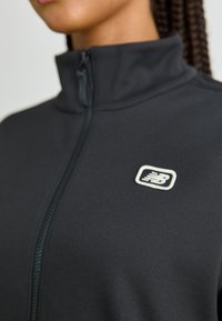 Schwarze Zip-Jacke mit Stehkragen, strukturiertem Stoff und einem weißen Logo-Akzent auf der linken Seite.