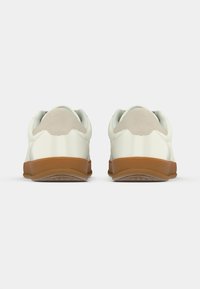 Ζευγάρι λευκών αθλητικών παπουτσιών με καφέ λαστιχένιες σόλες, εμφανιζόμενα από τη φτέρνα, σε απλό λευκό φόντο.