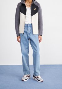 Nike mikina na zip v šedé a krémové barvě, světle modré džíny a bílé tenisky s tmavými detaily. Hladká látka, běžný střih a viditelné detaily loga.