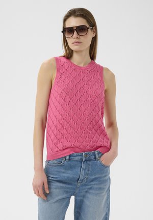 Vrouw draagt oversized bruine zonnebril, een mouwloos roze gebreid topje met diamantpatronen en een blauwe spijkerbroek, terwijl ze tegen een effen achtergrond staat.