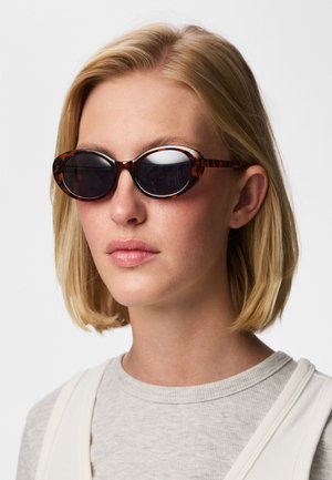 Lyhytukkainen nainen, jolla on soikeat kilpikonnakuosiset aurinkolasit ja vaaleanharmaa paita, valkoista taustaa vasten.