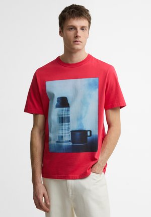Jonge man met een rood T-shirt met een blauw getinte afbeelding van een geruite thermosfles en een zwarte beker, gecombineerd met een witte broek, staand met één hand in de broekzak.