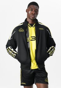 Sportswear set featuring a black zip-up jacket with yellow and white accents, a short yellow jersey, and black shorts. Textured material and logos visible.