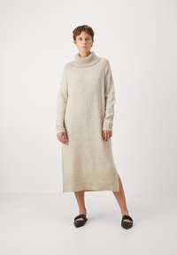 Abito in maglia beige con colletto alto, maniche lunghe, polsini e orlo a costine, dotato di uno spacco laterale per facilitare i movimenti.