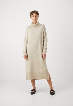 Beige strikket kjole med høyt krage, lange ermer, ribbestrikkede mansjetter og bærestykke, med en sideslisse for lettere bevegelse.