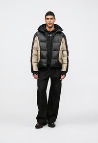 Casaco acolchoado com exterior preto e seções bege, detalhes vermelhos e capuz, usado por cima de um sweatshirt preto e calças largas escuras.