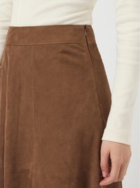 Detalhe de uma saia de camurça castanha com um fecho lateral, usada com uma blusa de manga longa bege com copa estriada, mostrando parte de uma mão e braço.