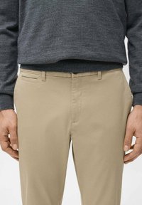 Mężczyzna w beżowych spodniach i ciemnoszarym swetrze, z rękami swobodnie opuszczonymi wzdłuż ciała, stojący na tle jednolitego jasnego tła.