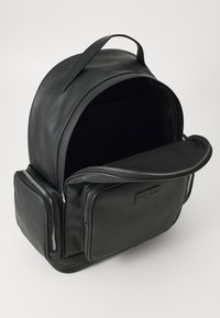 Svart lær ryggsekk med avrundet topp, glidelås lommer foran og en glatt tekstur. Åpen innside viser et minimalistisk design.