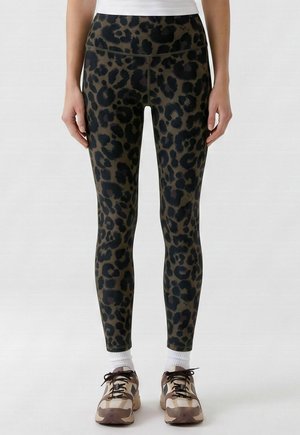 Leggings avec imprimé léopard vert foncé et noir portés avec des chaussettes blanches et des baskets camouflage marron sur une personne debout.